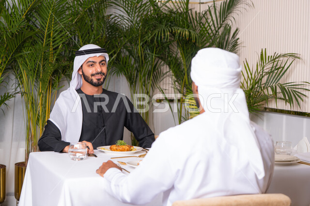 بناء علاقات اجتماعية ناجحة, تبادل الأحاديث والاخبار, مشاركة الأصدقاء الأوقات الممتعة, الاستمتاع بتناول الغداء في المطعم, صديقان عربيان خليجيان اماراتيان يرتديان الزي الاماراتي يجلسان على المائدة في إحدى المطاعم في الإمارات ويقومون بتناول الطعام