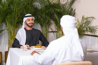 بناء علاقات اجتماعية ناجحة, تبادل الأحاديث والاخبار, مشاركة الأصدقاء الأوقات الممتعة, الاستمتاع بتناول الغداء في المطعم, صديقان عربيان خليجيان اماراتيان يرتديان الزي الاماراتي يجلسان على المائدة في إحدى المطاعم في الإمارات ويقومون بتناول الطعام