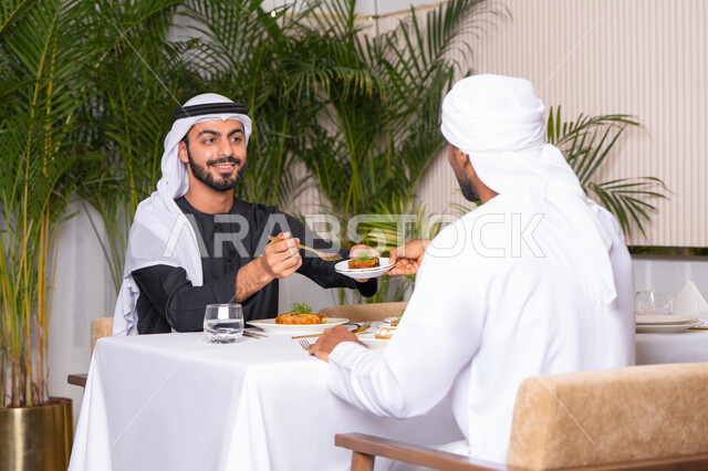 تبادل الأحاديث والاخبار, مشاركة الأصدقاء الأوقات الممتعة, الاستمتاع بتناول الغداء في المطعم, بناء علاقات اجتماعية ناجحة, صديقان عربيان خليجيان اماراتيان يرتديان الزي الاماراتي يجلسان على المائدة في إحدى المطاعم في الإمارات ويقومون بتناول الطعام