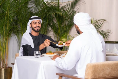 تبادل الأحاديث والاخبار, مشاركة الأصدقاء الأوقات الممتعة, الاستمتاع بتناول الغداء في المطعم, بناء علاقات اجتماعية ناجحة, صديقان عربيان خليجيان اماراتيان يرتديان الزي الاماراتي يجلسان على المائدة في إحدى المطاعم في الإمارات ويقومون بتناول الطعام