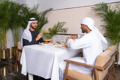 Building successful social relationships, exchanging conversations and news, sharing fun times with friends, enjoying lunch at a restaurant, two Emirati Gulf Arab friends wearing Emirati clothing sitting at a table in a restaurant in the Emirates and eating