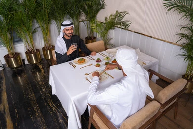 بناء علاقات اجتماعية ناجحة, تبادل الأحاديث والاخبار, مشاركة الأصدقاء الأوقات الممتعة, الاستمتاع بتناول الغداء في المطعم, صديقان عربيان خليجيان اماراتيان يرتديان الزي الاماراتي يجلسان على المائدة في إحدى المطاعم في الإمارات ويقومون بتناول الطعام