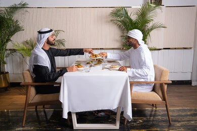 تبادل الأحاديث والاخبار, مشاركة الأصدقاء الأوقات الممتعة, الاستمتاع بتناول الغداء في المطعم, بناء علاقات اجتماعية ناجحة, صديقان عربيان خليجيان اماراتيان يرتديان الزي الاماراتي يجلسان على المائدة في إحدى المطاعم في الإمارات ويقومون بتناول الطعام