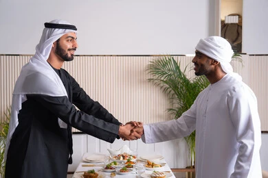 ايماءات السعادة والسرور، مشاركة الأصدقاء الأوقات الممتعة, بناء علاقات اجتماعية ناجحة, تبادل الأحاديث والاخبار, صديقان عربيان خليجيان اماراتيان يرتديان الزي الاماراتي يقومان بمصافحة بعضهما في إحدى المطاعم في الإمارات