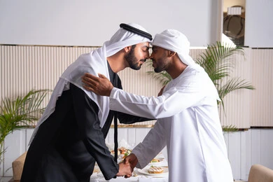 عقد صفقات عمل ناجحة والإتفاق على المشاريع الجديدة، المصافحة بين الأصدقاء في مطعم راقي في الإمارات العربية المتحدة، رجلان أعمال عربيان خليجيان إماراتيان يرتديان الغترة والكندورة الإماراتية ويقومان بقبلة الأنف التقليدية، التحية الإماراتية التقليدية