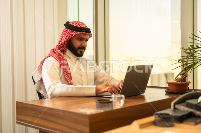 ايماءات تدل على التركيز لتحقيق أهداف الشركة، مفهوم خدمة العملاء، دمج العمل مع التقنية والتكنولوجيا, موظف عربي خليجي سعودي يجلس على كرسي مريح يرتدي الشماغ والثوب التقليدي يقوم بانهاء المهام اليومسة في مكتب شركة شعودية،بيئة عمل مريحة في الشركات والمكاتب