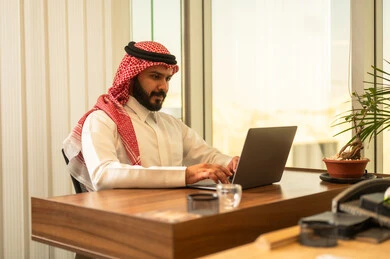 ايماءات تدل على التركيز لتحقيق أهداف الشركة، مفهوم خدمة العملاء، دمج العمل مع التقنية والتكنولوجيا, موظف عربي خليجي سعودي يجلس على كرسي مريح يرتدي الشماغ والثوب التقليدي يقوم بانهاء المهام اليومسة في مكتب شركة شعودية،بيئة عمل مريحة في الشركات والمكاتب