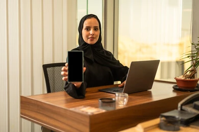 عرض شاشة سوداء على الهاتف المحمول، مفهوم خدمة العملاء، دمج العمل مع التقنية والتكنولوجيا, موظفة عربية خليجية سعودية تجلس على كرسي مريح ترتدي الحجاب والعباءة السوداء يقوم بانهاء المهام اليومسة في مكتب شركة شعودية،بيئة عمل مريحة في الشركات والمكاتب
