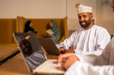 Professions and office work in the Sultanate of Oman, sending electronic mail via the computer, using modern and advanced technologies in work, an Arab Gulf Omani man in traditional attire sitting inside the office and using a laptop, communicating with clients remotely.