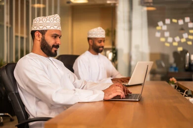 Sending electronic mail via the computer, using modern and advanced technologies in work, professions and office jobs in the Sultanate of Oman, an Arab Gulf Omani man in traditional attire sitting inside the office and using a laptop, communicating with clients remotely.
