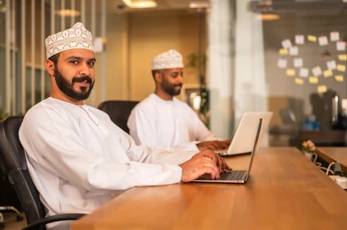 Professions and office work in the Sultanate of Oman, sending electronic mail via the computer, using modern and advanced technologies in work, an Arab Gulf Omani man in traditional attire sitting inside the office and using a laptop, communicating with clients remotely.