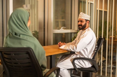 The concept of dialogue and exchanging opinions in business achievement plans, administrative and office jobs in the UAE, conducting hiring and employment interviews, an Arab Gulf Omani manager wearing a dishdasha and kumma inside the office conducting a job interview with a female employee, equality between men and women.