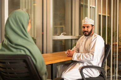 مهن ووظائف إدارية مكتبية في الإمارات، مفهوم التحاور وتبادل الأراء في خطط إنجاز الأعمال، إجراء مقابلة التعيين والتوظيف، صورة مقربة لمدير عربي خلجي عماني يرتدي الدشداشة و الكمه  داخل المكتب يقوم بإجراء مقابلة عمل مع موظفة، المساواة بين المرأة والرجل