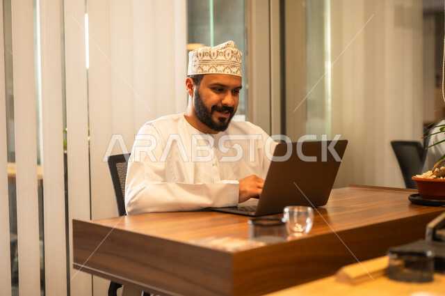 Omani office professions and jobs, the concept of business management and achieving company goals, an Arab Gulf Omani businessman wearing traditional attire sitting in his office working on a laptop with gestures of enthusiasm and happiness, accomplishing remote work tasks.