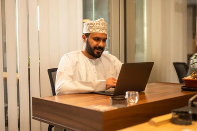 مهن ووظائف مكتبية عمانية ، مفهوم ادارة الاعمال وتحقيق اهداف الشركة،رجل اعمال عربي خليجي عماني يرتدي التقليدي يجلس في مكتبه يعمل على الحاسوب المحمول بايماءات الحماس والسعادة، انجاز مهام العمل عن بعد