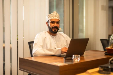 مهن ووظائف مكتبية عمانية ، مفهوم ادارة الاعمال وتحقيق اهداف الشركة،رجل اعمال عربي خليجي عماني يرتدي التقليدي يجلس في مكتبه يعمل على الحاسوب المحمول بايماءات الحماس والسعادة، انجاز مهام العمل عن بعد