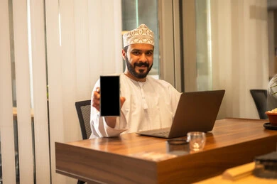 Using a mobile phone to manage business affairs online, online administrative office jobs, integrating work with technological technologies, the concept of remote work, an Arab Gulf Omani man wearing traditional attire sitting behind a desk, a black screen with a mobile phone.