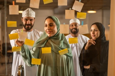 أوراق الملاحظات الملونة المعلقة, بيئة العمل في الشركات العمانية ،كتابة وتدوين شيء ما , مهن وأعمال مكتبية, صورة لمجموعة موظفين عربيين خليجيين عمانيين يقومون  بلصق اوراق  ملاحظة على واجهة زجاجية داخل المكتب
