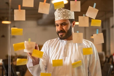 Writing and noting something, hanging colored sticky notes, the work environment in Omani companies, office professions and jobs, a close-up image of an Omani Gulf Arab man sticking a note on a glass surface inside the office.