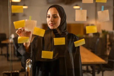 Writing and noting something, hanging colorful sticky notes, the work environment in Omani companies, office professions and jobs, a close-up image of an Omani Gulf Arab woman sticking a note on a glass surface inside the office.