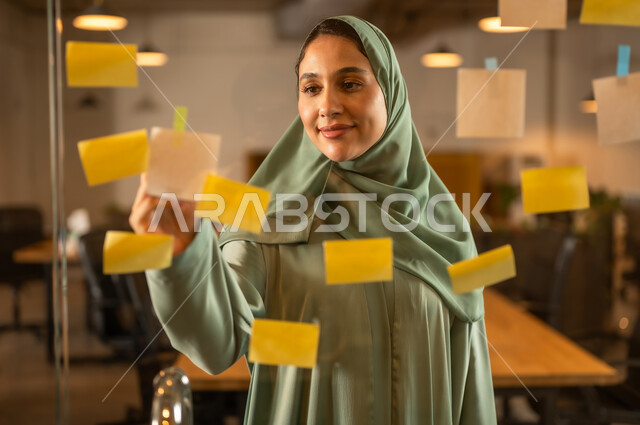 بيئة العمل في الشركات عمانية , مهن وأعمال مكتبية,كتابة وتدوين شيء ما, أوراق الملاحظات الملونة المعلقة,  صورة مقربة لامرأة عربية خليجية عمانية  تقوم بلصق ورقة ملاحظة على واجهة زجاجية داخل المكتب