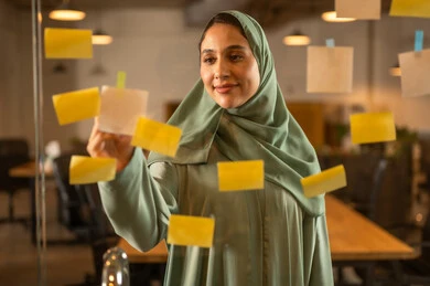 The work environment in Omani companies, professions and office work, writing and noting something, hanging colored sticky notes, a close-up image of an Omani Gulf Arab woman sticking a note on a glass surface inside the office.
