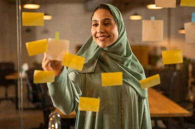 Writing and noting something, hanging colorful sticky notes, the work environment in Omani companies, office professions and jobs, a close-up image of an Omani Gulf Arab woman sticking a note on a glass surface inside the office.