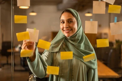 The work environment in Omani companies, professions and office work, writing and noting something, hanging colored sticky notes, a close-up image of an Omani Gulf Arab woman sticking a note on a glass surface inside the office.