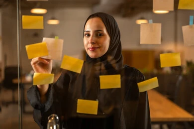 Writing and noting something, hanging colorful sticky notes, the work environment in Omani companies, office professions and jobs, a close-up image of an Omani Gulf Arab woman sticking a note on a glass surface inside the office.