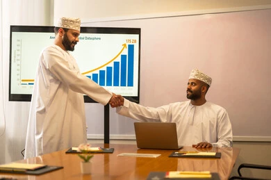 Successful new business deals between businessmen, conducting a job interview, two Omani Arab Gulf men wearing traditional attire shaking hands, expanding business fields, partnership in business and engineering projects, studying statistical data, analyzing charts.