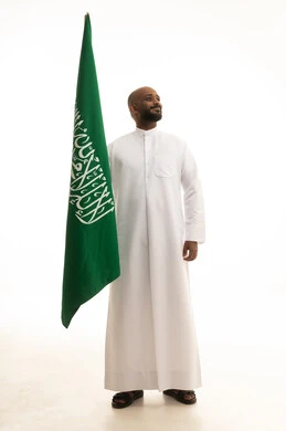 Flag Day is March 11, the celebration of the National Day is on the 8th of Rabi' al-Awwal, pride and honor in belonging to the Kingdom, the anniversary of the Saudi National Day, a portrait of a smiling Arab Saudi Gulf man wearing traditional attire holding the Saudi flag in his hand, white background.