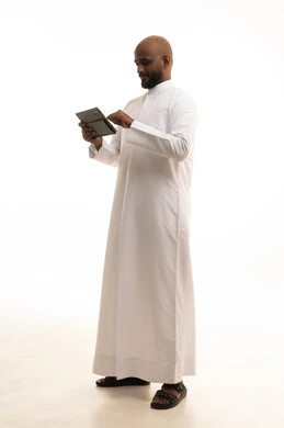 Using a modern and advanced technical device, chatting with friends and relatives via the tablet, gestures of joy and happiness, an Arab Gulf Saudi man wearing traditional attire holding the tablet in his hand, white background.