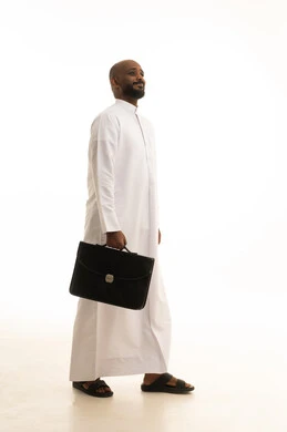 مفهوم ادارة الاعمال والمشاريع، المشي والذهاب الى مكان ما، الاهتمام بالمظهر الخارجي والأناقة، بورتريه لرجل أعمال عربي خليجي سعودي مبتسم يرتدي الثوب التقليدي يحمل بيده حقيبة جلد سوداء ينظر  بإيماءات البشاشة والسرور، صورة الجسم بالطول الكامل، خلفية بيضاء