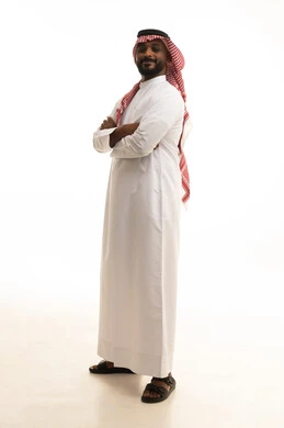 الوقوف بايدي متقاطعه ،  مفهوم الرجولة والثقة بالنفس، الاهتمام بالمظهر الخارجي والأناقة، بورتريه لرجل عربي خليجي سعودي يرتدي الثوب التقليدي والشماغ  ينظر بتعابير السرور، صورة الجسم بالطول الكامل، خلفية بيضاء