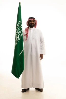 بورتريه لرجل عربي سعودي خليجي مبتسم يرتدي الشماغ والثوب التقليدي يحمل بيده علم السعودية , الفخر و الاعتزاز بالانتماء للمملكة, يوم العلم 11مارس، الاحتفال باليوم الوطني 8 ربيع الأول, ذكرى العيد الوطني السعودي,خلفية بيضاء
