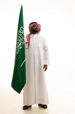 Flag Day is March 11, the celebration of the National Day on the 8th of Rabi' al-Awwal, pride and honor in belonging to the Kingdom, the anniversary of the Saudi National Day, a portrait of a smiling Arab Saudi Gulf man wearing a ghutrah and traditional thobe holding the Saudi flag in his hand, with a white background.