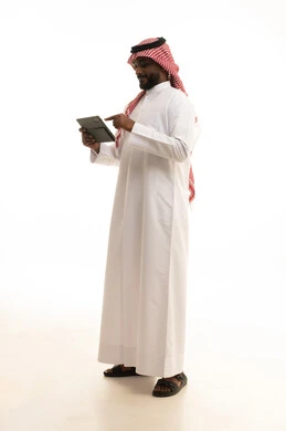 استخدام جهاز تقني حديث ومتطور، الدردشة مع الأصدقاء والأقارب عبر التابلت، ايماءات الفرح والسعادة، رجل عربي خليجي سعودي يرتدي الثوب التقليدي والشماغ يمسك بيده الجهاز اللوحي، خلفية بيضاء