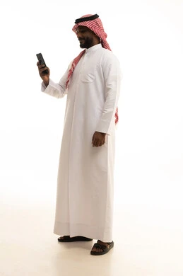 اجراء بث مباشر، استخدام جهاز تقني حديث ومتطور، الدردشة مع الأصدقاء والأقارب عبر الجوال، ايماءات الفرح والسعادة، رجل عربي خليجي سعودي يرتدي الثوب التقليدي والشماغ يمسك بيده الهاتف المحمول، خلفية بيضاء