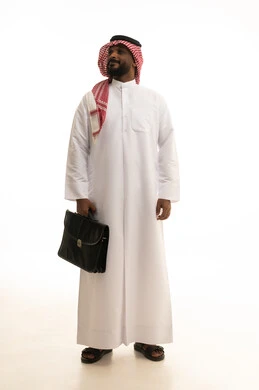 الاهتمام بالمظهر الخارجي والأناقة، مفهوم ادارة الاعمال والمشاريع، المشي والذهاب الى مكان ما، بورتريه لرجل أعمال عربي خليجي سعودي مبتسم يرتدي الثوب التقليدي والشماغ يحمل بيده حقيبة جلد سوداء ينظر إلى الكاميرا بإيماءات البشاشة والسرور، صورة الجسم بالطول الكامل، خلفية بيضاء