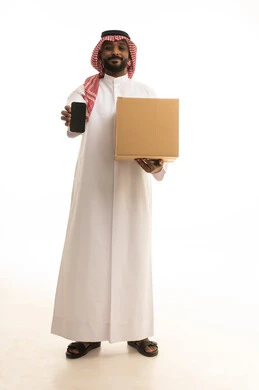 Integrating modern technologies into daily life, reliable shipping and delivery processes, fast delivery services, ordering and receiving delivery, a portrait of an Arab Saudi Gulf man wearing traditional attire and a ghutrah holding a mobile phone working in delivering orders and goods to customers, online shopping and online orders, white background.