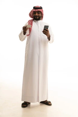 The evolution of technology and technical devices, the concept of remote payment and settlement through mobile applications, the use of electronic bank cards, a portrait of a Saudi Arabian Gulf Arab man wearing traditional attire and a ghutrah using a mobile phone and holding a white card, white background.