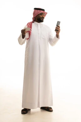The use of electronic bank cards, the concept of remote payment and settlement through mobile applications, the evolution of technology and technical devices, a portrait of an Arab Gulf Saudi man wearing traditional attire and a ghutrah using a mobile phone and holding a white card, white background.