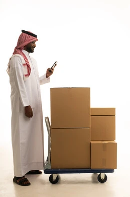 Integrating modern technologies into daily life, organizing and arranging the home, cardboard boxes and containers, a portrait of a Saudi Arabian Gulf man wearing traditional attire and a ghutrah, carrying boxes of household items while holding a mobile phone, owning a new home and family stability, modern housing and ownership, full-body image, white background.