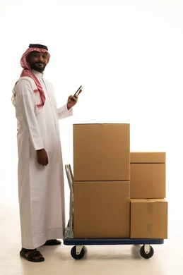 Owning a new home and family stability, integrating modern technologies into daily life, organizing and arranging the home, cardboard boxes and containers, a portrait of a Saudi Arabian Gulf man wearing traditional attire and a ghutrah carrying household items boxes while holding a mobile phone, modern housing and ownership, full-body image, white background.