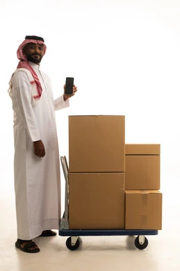 Integrating modern technologies into daily life, organizing and arranging the home, cardboard boxes and containers, a portrait of a Saudi Arabian Gulf man wearing traditional attire and a ghutrah, carrying boxes of household items while holding a mobile phone, owning a new home and family stability, modern housing and ownership, full-body image, white background.