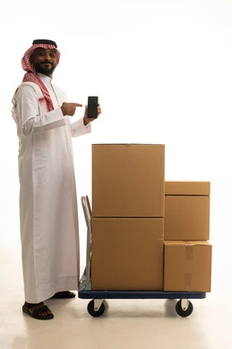 استخدام التكنولوجيا الحديثة ، ترتيب وتنظيم المنزل، صناديق وحاويات كرتونية، بورتريه لرجل عربي خليجي سعودي يرتدي الزى التقليدي والشماغ يقوم بنقل صناديق الأغراض المنزلية يمسك بيده الجوال، امتلاك سكن جديد والاستقرار العائلي، اسكان وتمليك حديث ، صورة الجسم بالطول الكامل، خلفية بيضاء