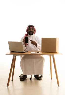 عرض شاشة سوداء فارغة، تصفح وسائل التواصل الاجتماعي، إستخدام اجهزة تقنية حديثة ، العمل عن بعد ، مفهوم التسويق الإلكتروني وطلبات الاونلاين ،بورتريه لرجل عربي خليجي سعودي يرتدي الزى التقليدي والشماغ يستخدم جهاز الحاسوب المحمول يشير الى الهاتف المحمول امامه اكياس التسوق الورقية ،خلفية بيضاء