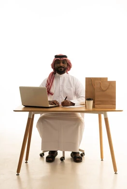 Taking important notes, the concept of electronic marketing and online orders, Saudi professions and jobs, a portrait of a Saudi Gulf Arab man wearing traditional attire and a ghutrah using a laptop with shopping bags in front of him, browsing social media, using modern technological devices, working remotely, white background. Taking important notes, the concept of electronic marketing and online orders, Saudi professions and jobs, a portrait of a Saudi Gulf Arab man wearing traditional attire and a ghutrah using a laptop with shopping bags in front of him, browsing social media, using modern technological devices, working remotely, white background.