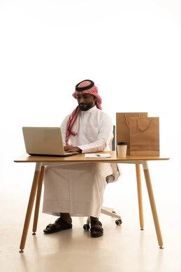 عقد إجتماع عمل عبر اللابتوب ، مفهوم التسويق الإلكتروني وطلبات الاونلاين ، مهن ووظائف سعودية ، بورتريه لرجل عربي خليجي سعودي يرتدي الزى التقليدي والشماغ يستخدم جهاز الحاسوب المحمول وامامه اكياس التسوق الورقية ، تصفح وسائل التواصل الاجتماعي، إستخدام اجهزة تقنية حديثة ، العمل عن بعد ،خلفية بيضاء