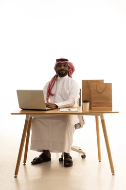 مفهوم التسويق الإلكتروني وطلبات الاونلاين ، عقد إجتماع عمل عبر اللابتوب ، مهن ووظائف سعودية ، بورتريه لرجل عربي خليجي سعودي يرتدي الزى التقليدي والشماغ يستخدم جهاز الحاسوب المحمول وامامه اكياس التسوق الورقية ، تصفح وسائل التواصل الاجتماعي، إستخدام اجهزة تقنية حديثة ، العمل عن بعد ،خلفية بيضاء